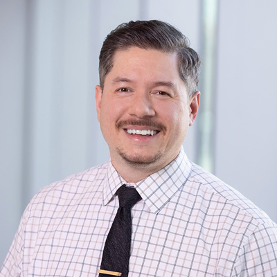 Alex Freedman smiling in a checkered shirt and dark tie, standing in front of a softly lit indoor background.