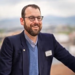 William Kubie wearing a blue dress shirt, blue sweater vest, and a blue sport coat. He is standing outside and he is smiling.