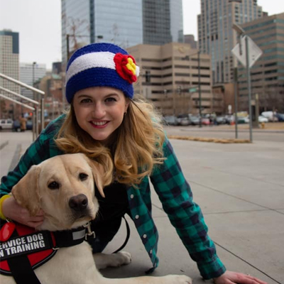 Elly Lewis smiling while kneeling next to a yellow lab wearing a red “Service Dog in Training” vest, with a city skyline in the background.