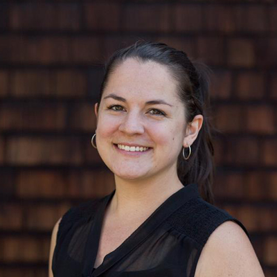 Haley Gray smiling wearing a black blouse and small gold hoop earrings, standing in front of brick wall.