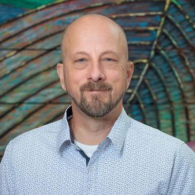 Paul Wedlake smiling wearing a dress shirt, standing in front of blue and green background