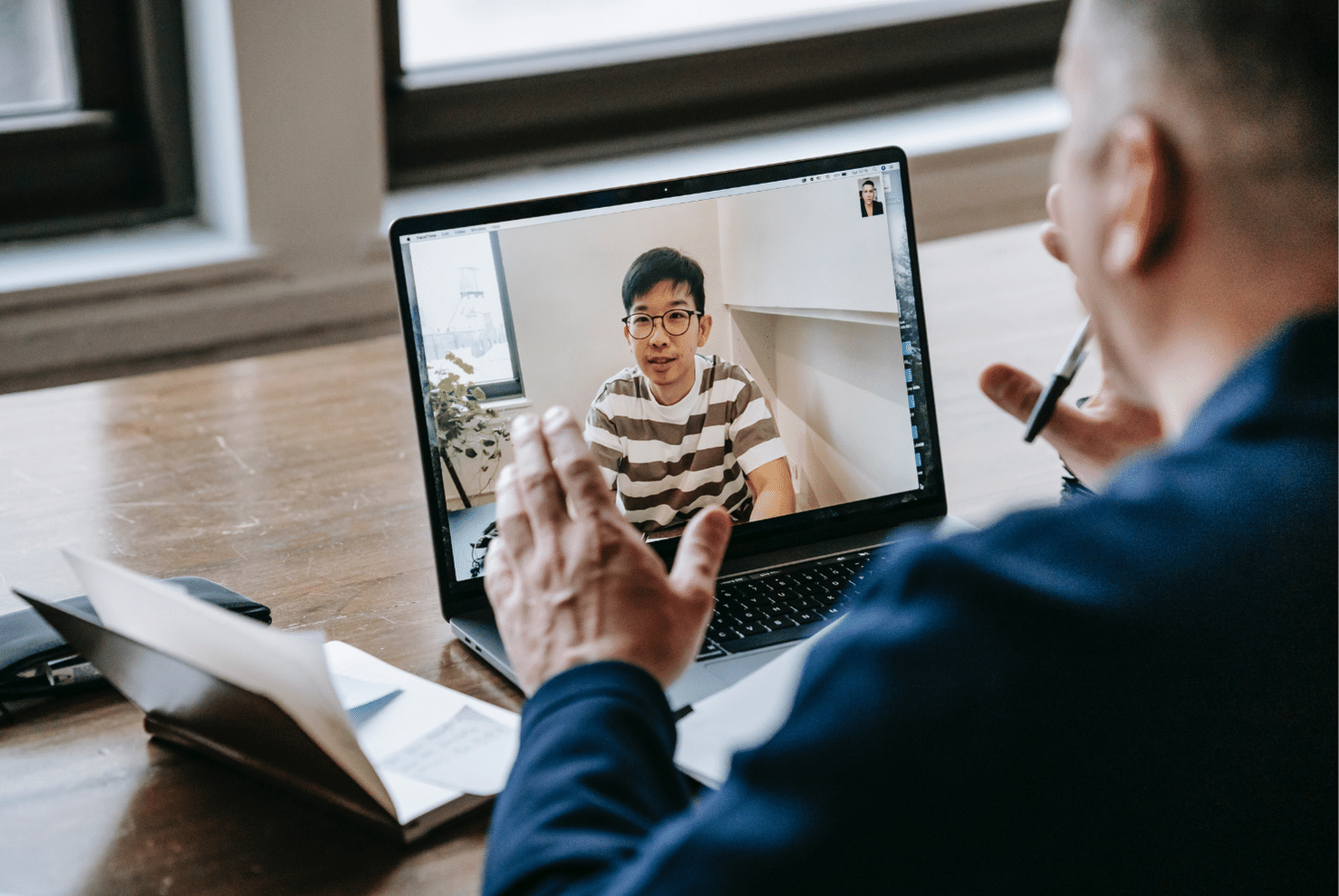 A man talking to someone through a video call on a laptop while a third person's face is shown in the top right corner of the screen.