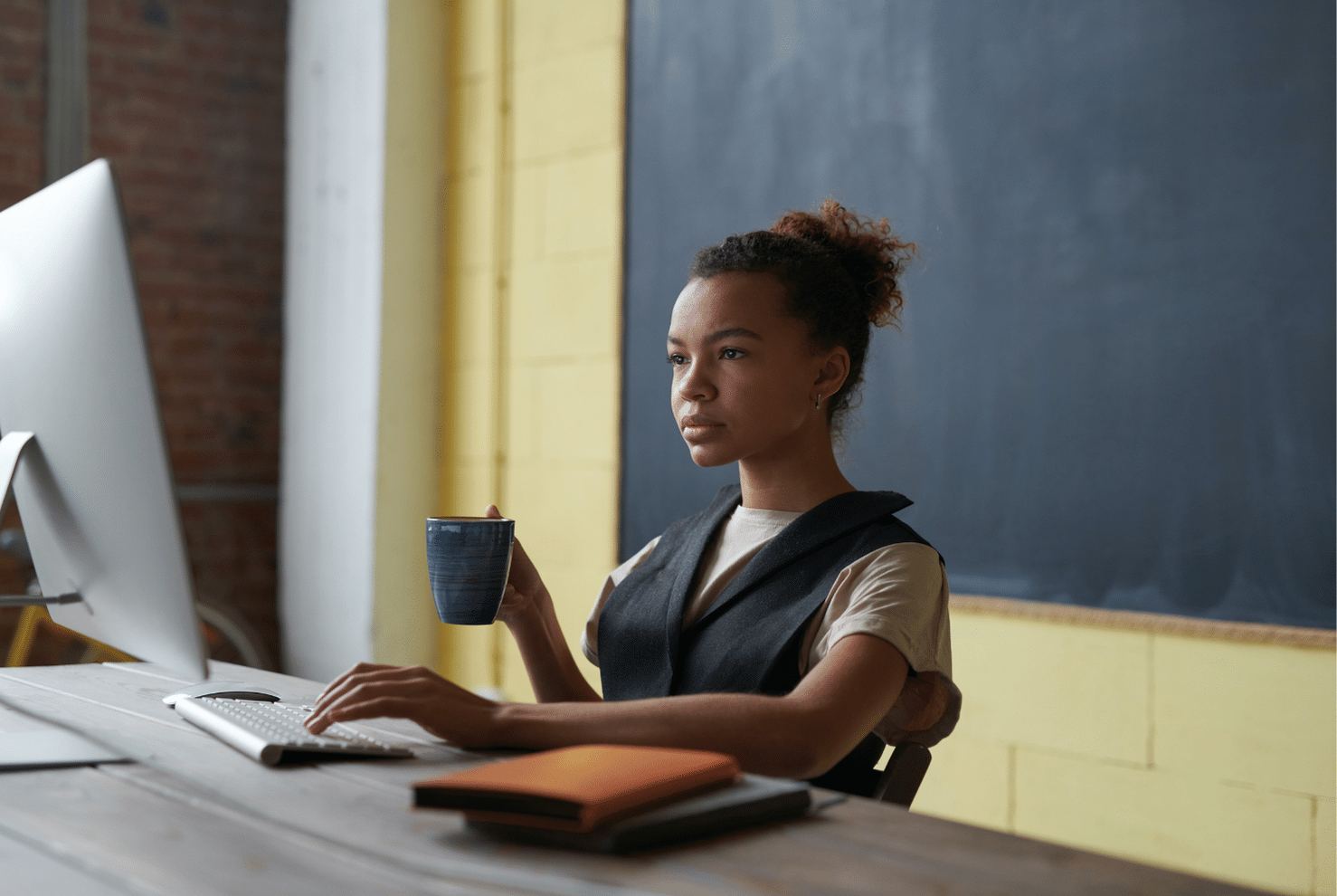 A woman sits down at her computer while holding a mug.