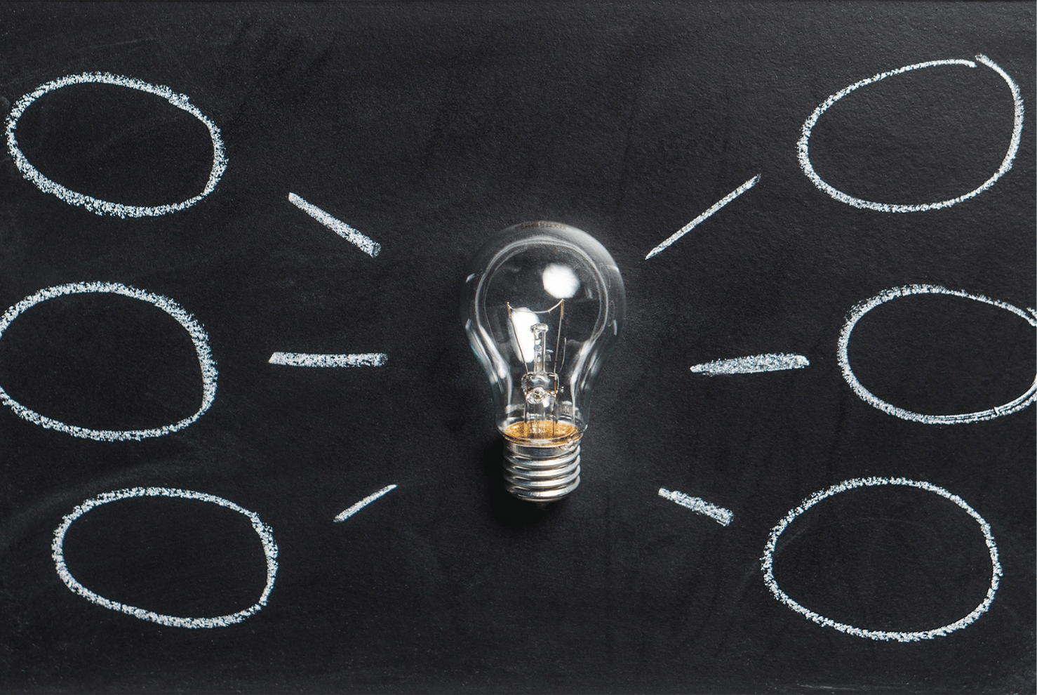 A single lightbulb laying on a blackboard with chalked lines and circles surrounding it.