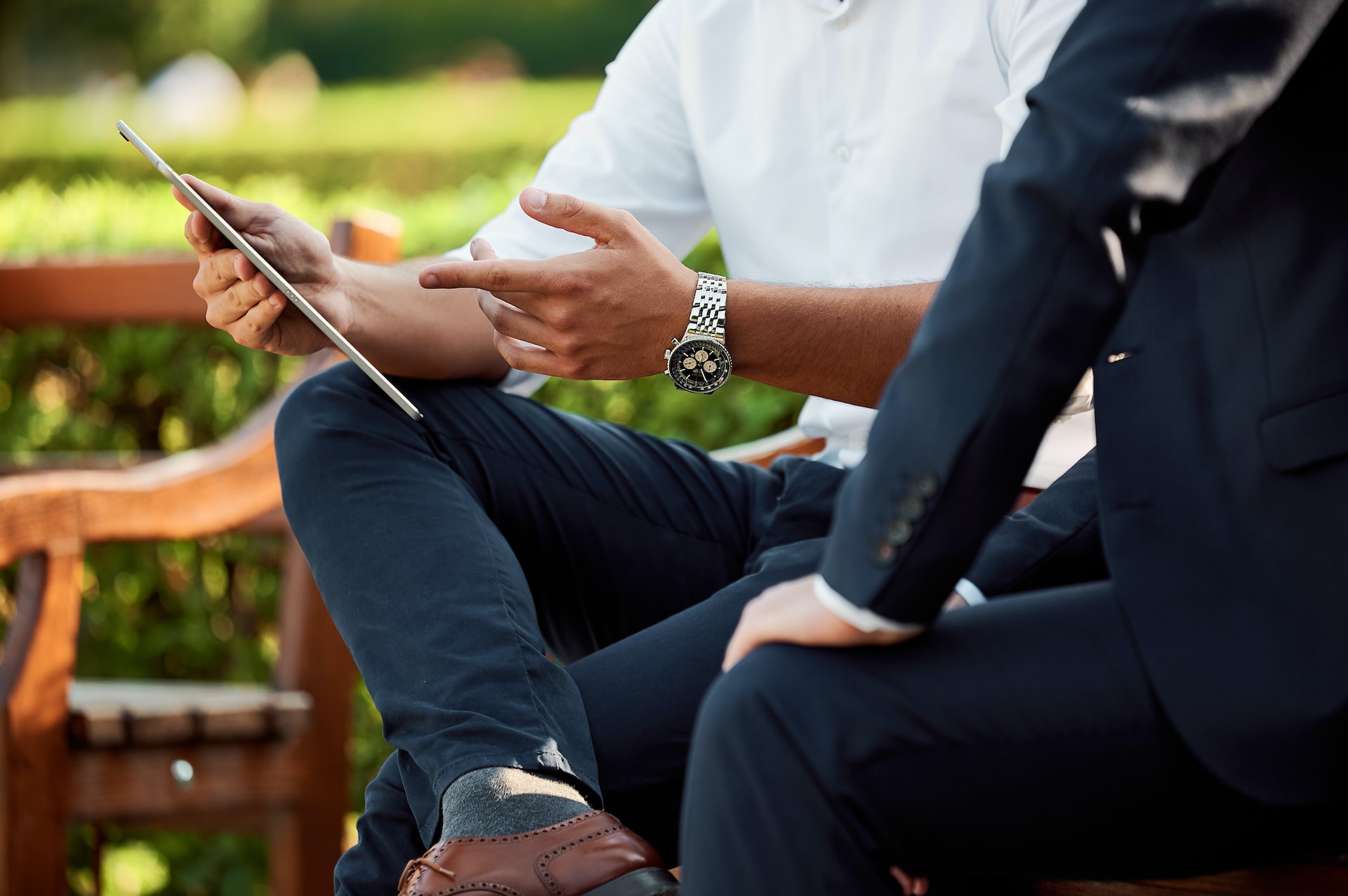 A close-up of a man's finger pointing at a tablet he is holding while crossing his legs and sitting beside another person.