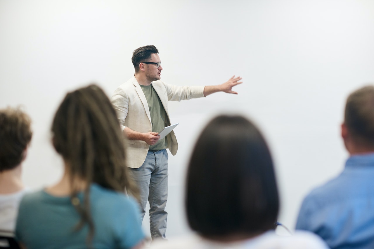 A man is lecturing and standing in front of students while holding a tablet and raising his arm towards a blank wall.