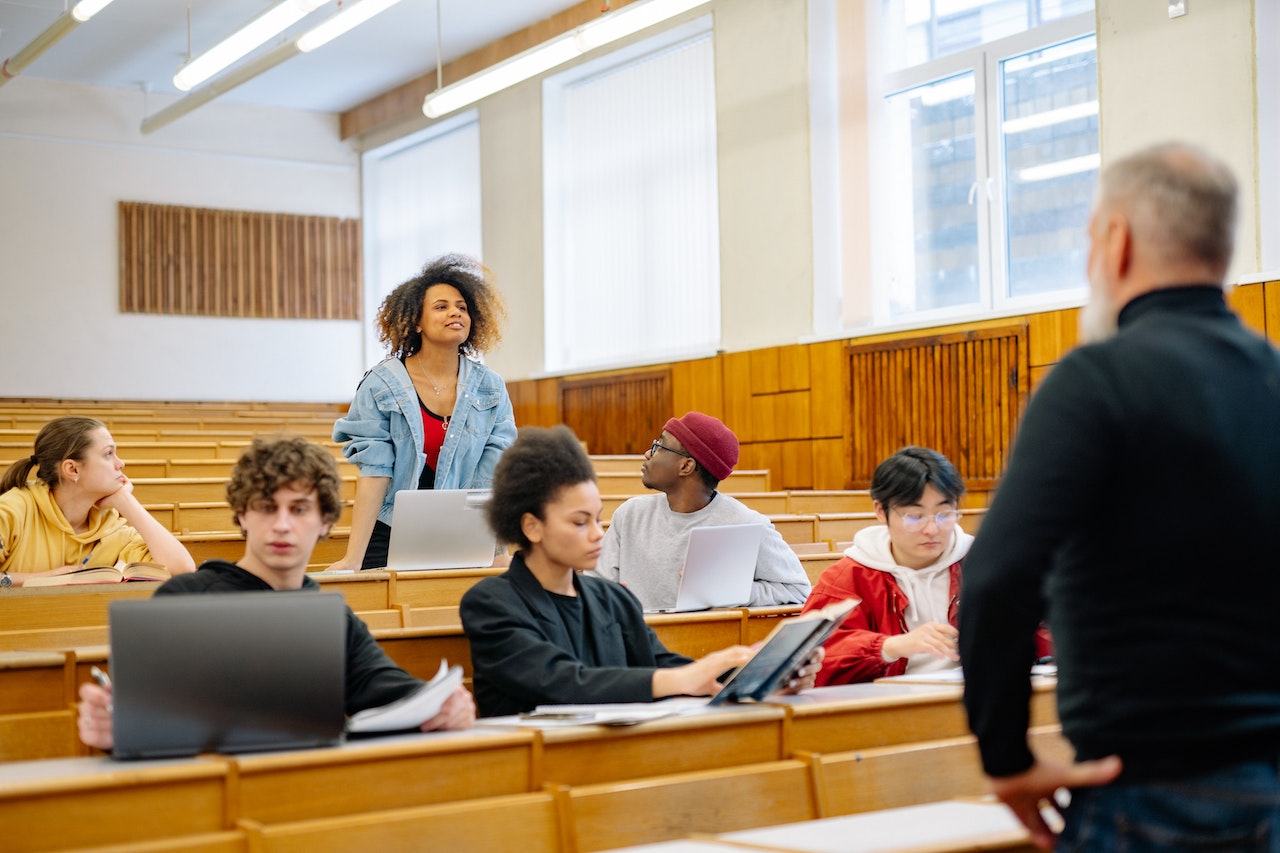 A female student in the back row is standing up to speak during a lecture while two students and the professor look at her.