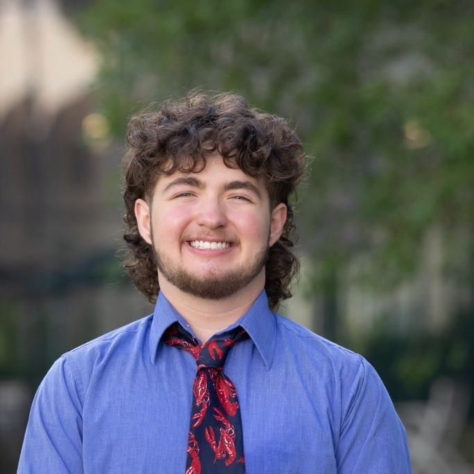 Professional photo of Cayson in front of trees. Cayson is a white man with short, curly, brown hair and a short brown beard. Cayson is wearing a long sleeve blue shirt that is buttoned up and a tie with a red crawfish pattern.