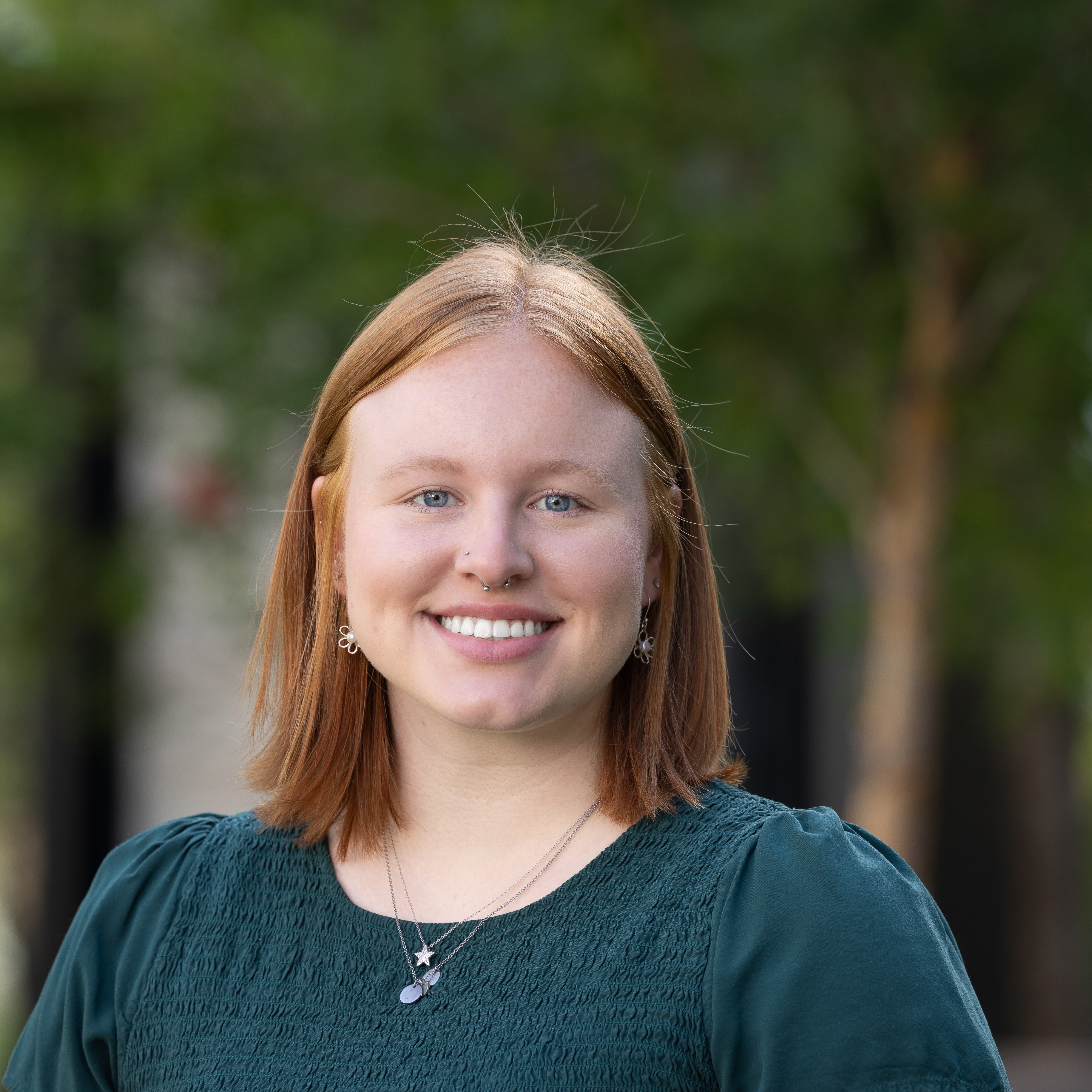 Professional photo of Katelyn in front of the trees. Katelyn is a white woman with shoulder-length red hair. Katelyn is wearing a dark green short shirt and three silver necklaces.
