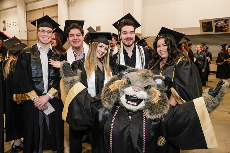 Five students and Milo the Lynx mascot in commencement regalia smiling in celebration.
