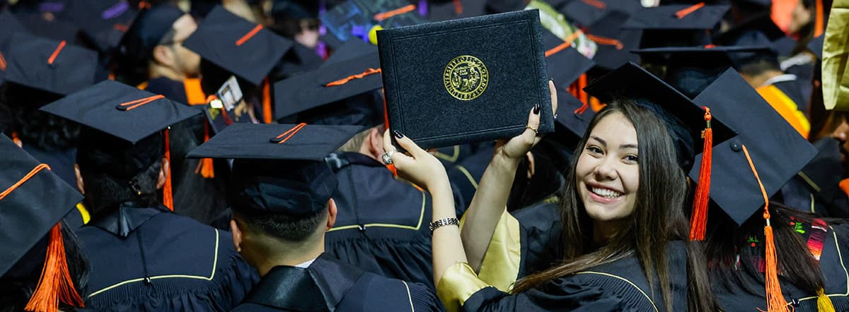 Graduate in commencement regalia proudly holding up a diploma cover in a crowd of graduates.