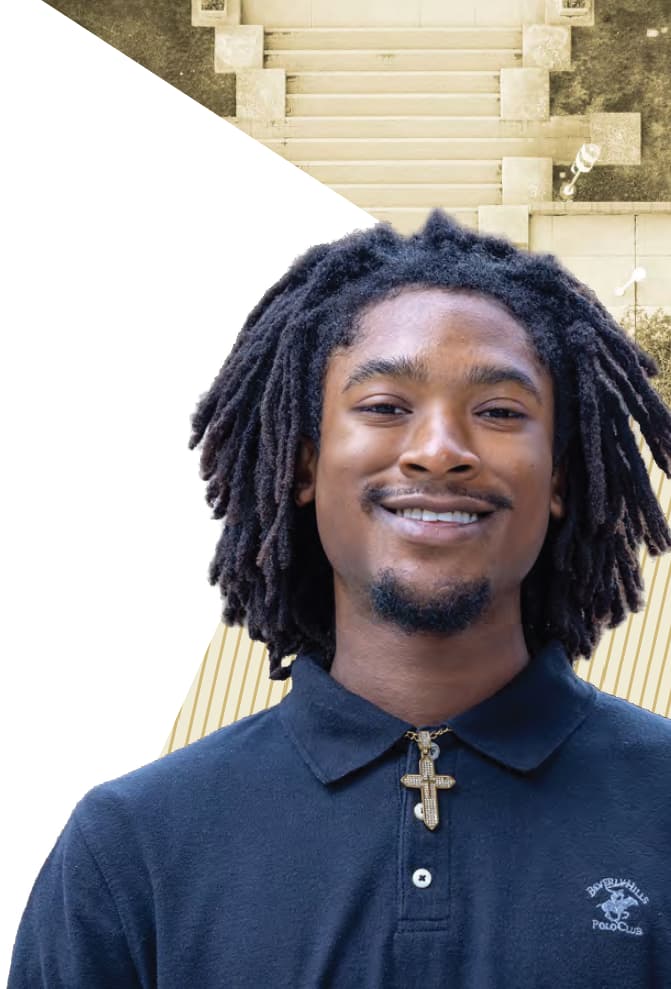 Man in a navy polo, facial hair, and black hair in locks smiling with his teeth.