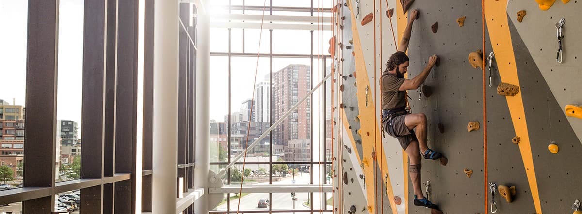 A person climbing a tall rock wall in a room of windows overlooking a city.