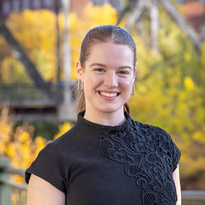 Woman in a black dress and hair pulled back smiling with her teeth. Fall leaves and a bridge are behind her.
