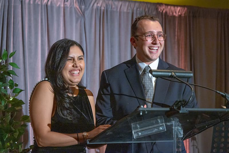 Two students at a podium speaking in professional attire, both are smiling with their teeth and mouths open. 