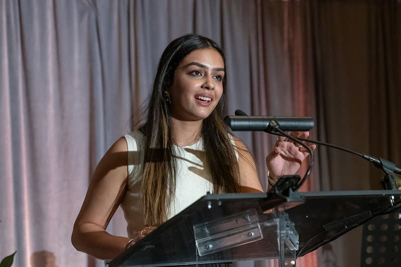 Woman with long dark hair wearing professional attire stands at a podium speaking. 