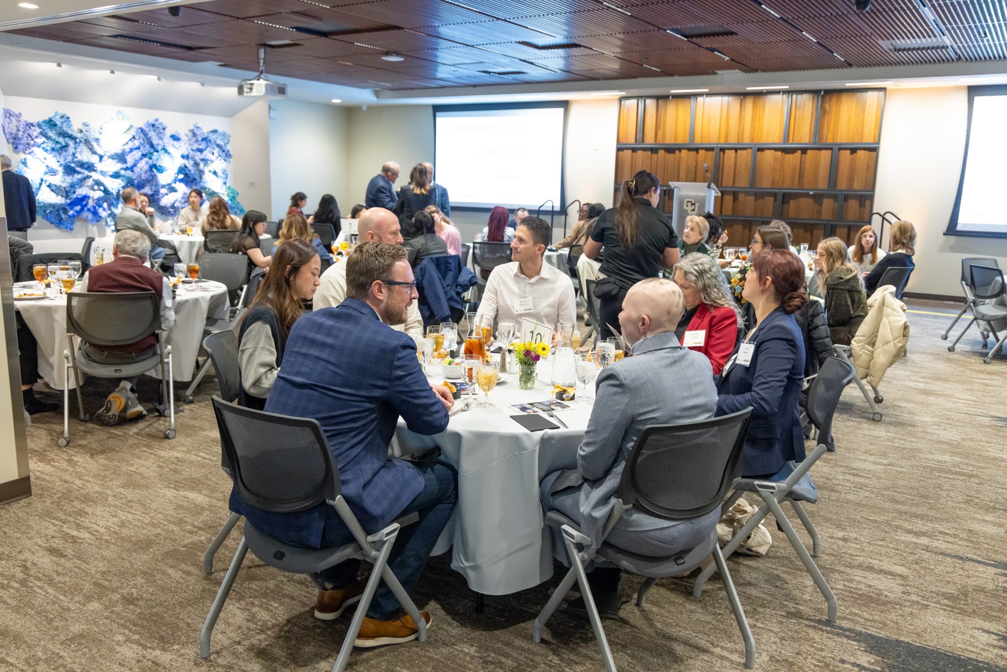 Wide angle of the whole room with people in business attire seated at round tables, art is visible in the background.