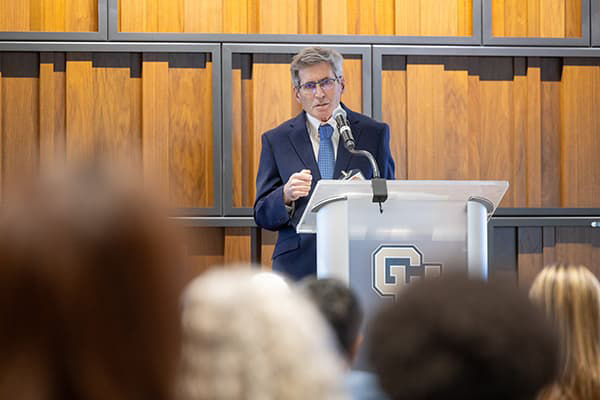 Bob Wolfson wearing a blue suite and tie speaking at a lectern with an interlocking CU mark on the font. 