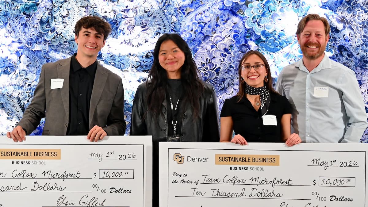 Four people in business attire smile while holding giant checks in front of an artistic background.