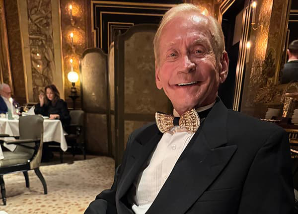 Man in a bow-tie and suite smiling proudly with his teeth seated in a fancy dining area.