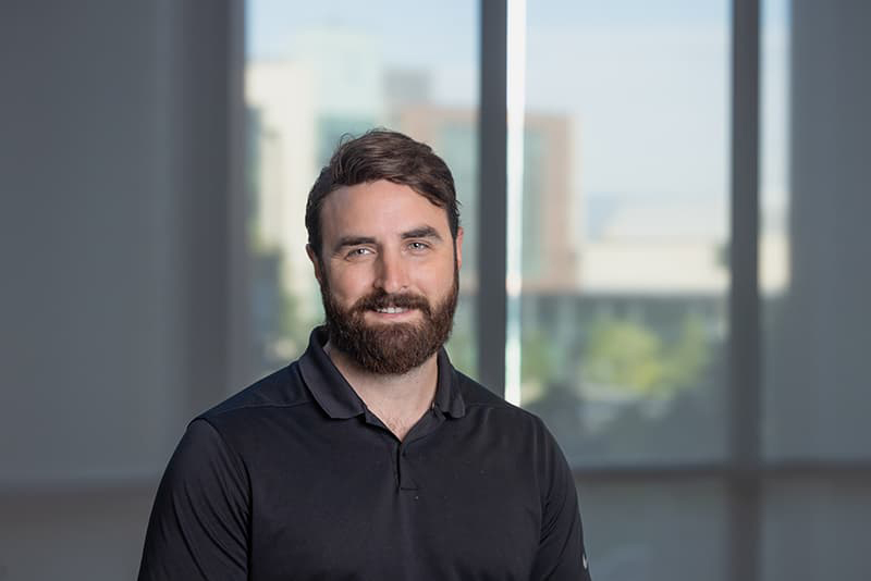 Headshot of Gentry Hewitt in a black polo, dark hair and beard, smiling with his teeth.
