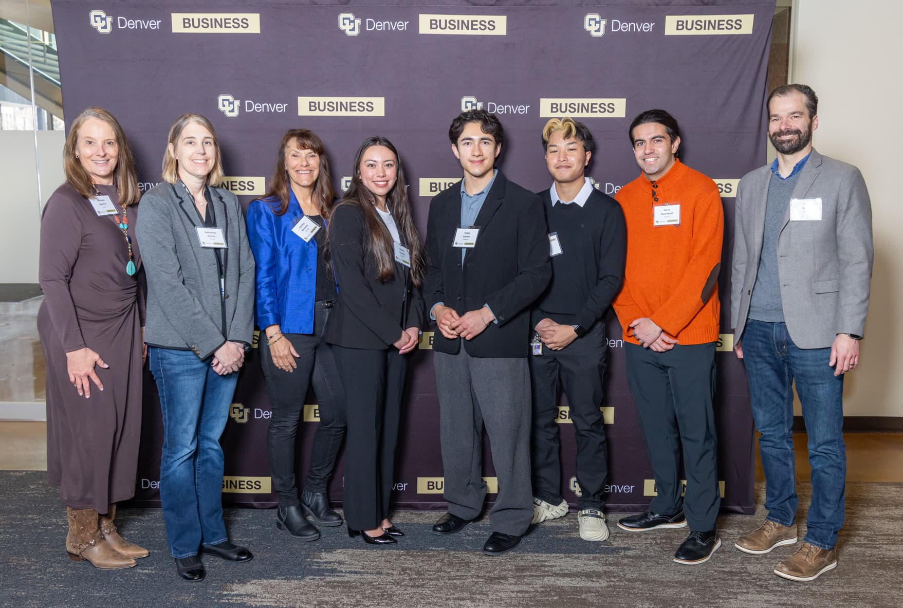 Eight people dressed in business casual line up smiling together against a CU Denver Business School backdrop.