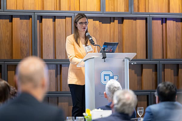 Katie Hughes in a yellow blazer speaks at a lectern with the gold interlocking CU on the front, 
