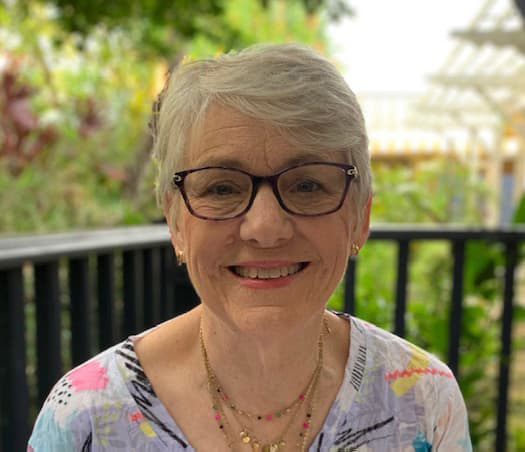 Woman with short white hair, dark-rimmed glasses, smiles with her teeth while sitting outside.