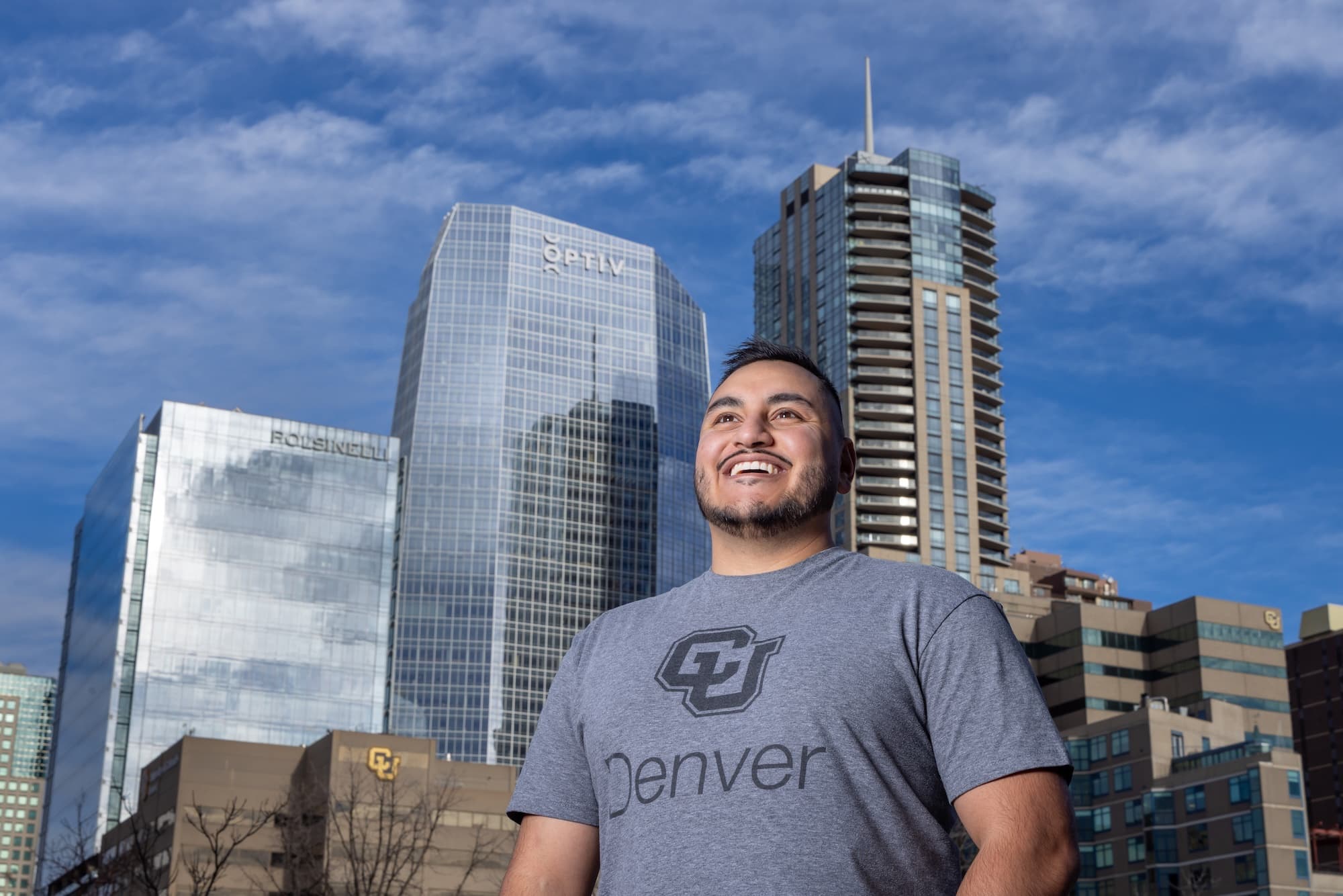 Man in a CU Denver branded gray shirt smiling with his teeth and the lower downtown Denver skyline is visible behind him.