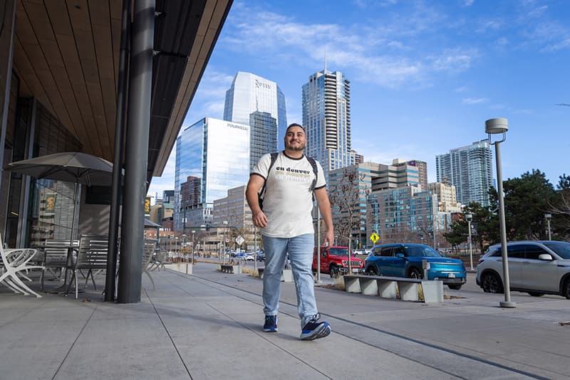 A man in a CU Denver branded shirt walks on campus with the lower downtown Denver skyline behind him. 