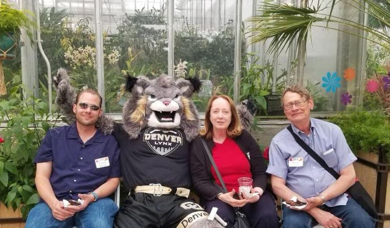 Three people seated with Milo the Lynx mascot surrounded by green plants.