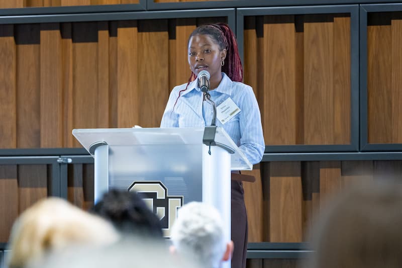 Woman in a blue collard blouse and long hair pulled into a pony tail speaks at a lectern. 