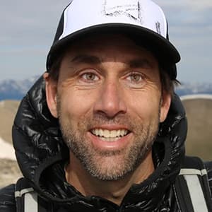 A man's face up close wearing a baseball cap and black hooded winter coat smiling with his teeth.