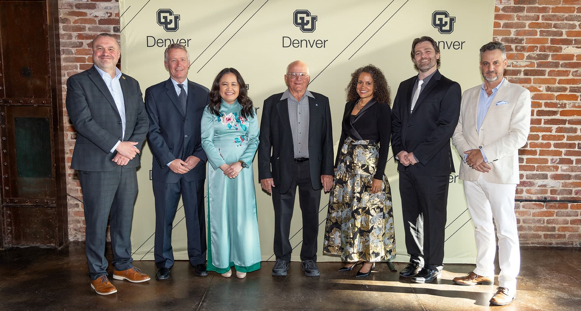 Seven people in business professional attire smile proudly in front of a CU Denver backdrop.