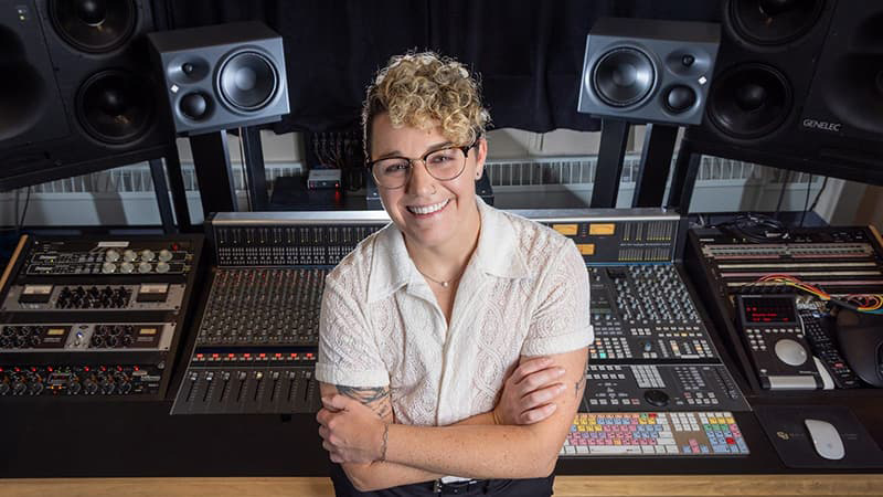 Person with short hair and glasses smiling with their teeth. Their arms are crossed and they lead against a technical board.