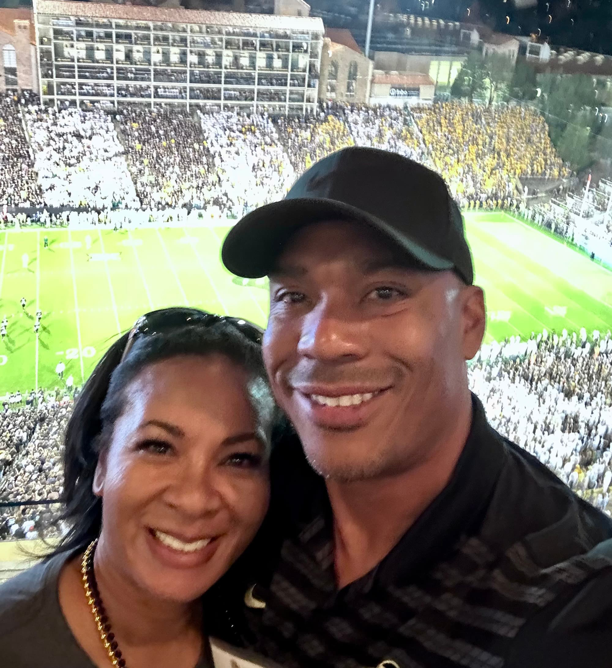 A male and female couple dressed in black taking a selfie with a football game in the background.