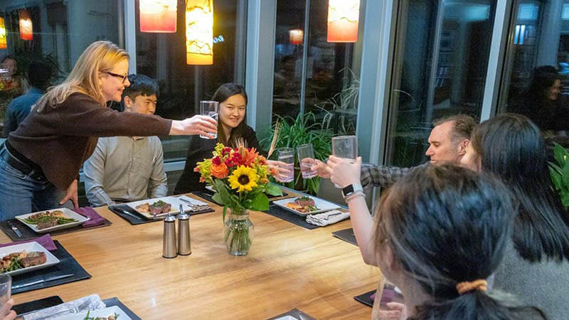 Group of people seated at a dinner table clinking glasses together in the center.