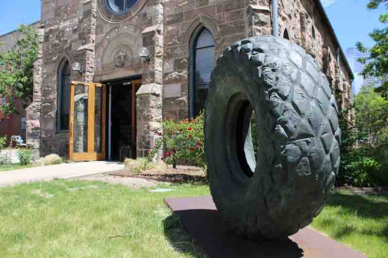A large bronze-cast tire sculpture sits upright in front of a historical church building made of gray stone. 