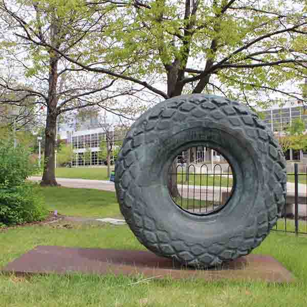 Large, bronze cast sculpture of a tire sitting upright on a platform in a grassy area.