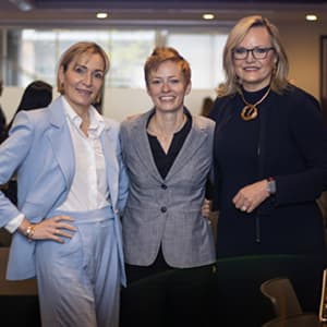 Three women in business professional attire pose together smiling.