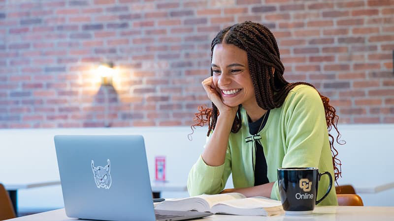 Woman seated and working on a laptop smiling. A brick wall is behind her.