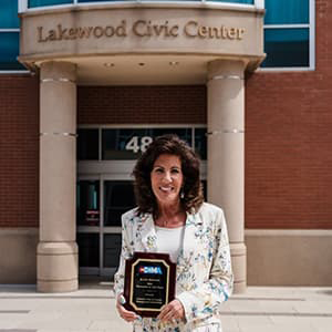 Kathy Hodgson in a white, floral suite proudly holding a plaque standing in front of the Lakewood Civic Center.