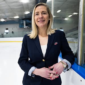 Woman in a dark suit jacket and long blonde hair smiles with her teeth while standing on a hockey rink.