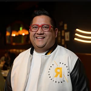 Man smiling with his teeth wearing red-framed glasses and a white letterman style jacket.