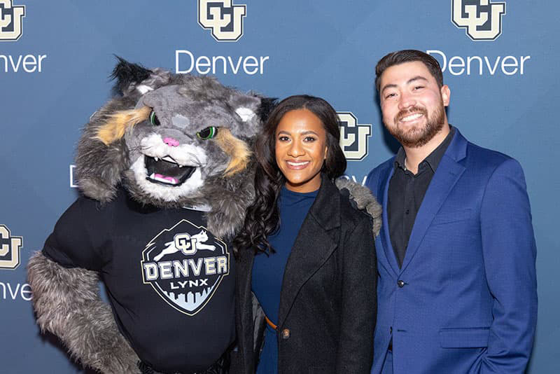 Two professionally dressed people pose with Milo the Lynx Mascot against a CU Denver branded backdrop.