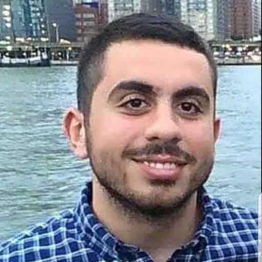 Taha Yahya with short dark hair smiling with his teeth wearing a blue plaid shirt.