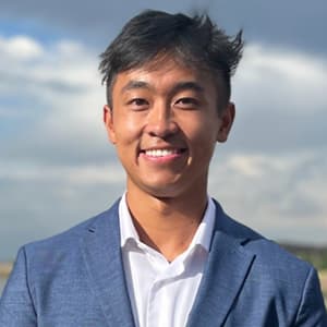 Man with dark hair wearing a blue suit jacket stands outside and smiles with his teeth.