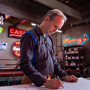 Man working on a sketch at a table with neon signs in the background.