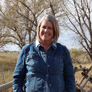 Woman in a denim jacket smiles with her teeth while standing outside.