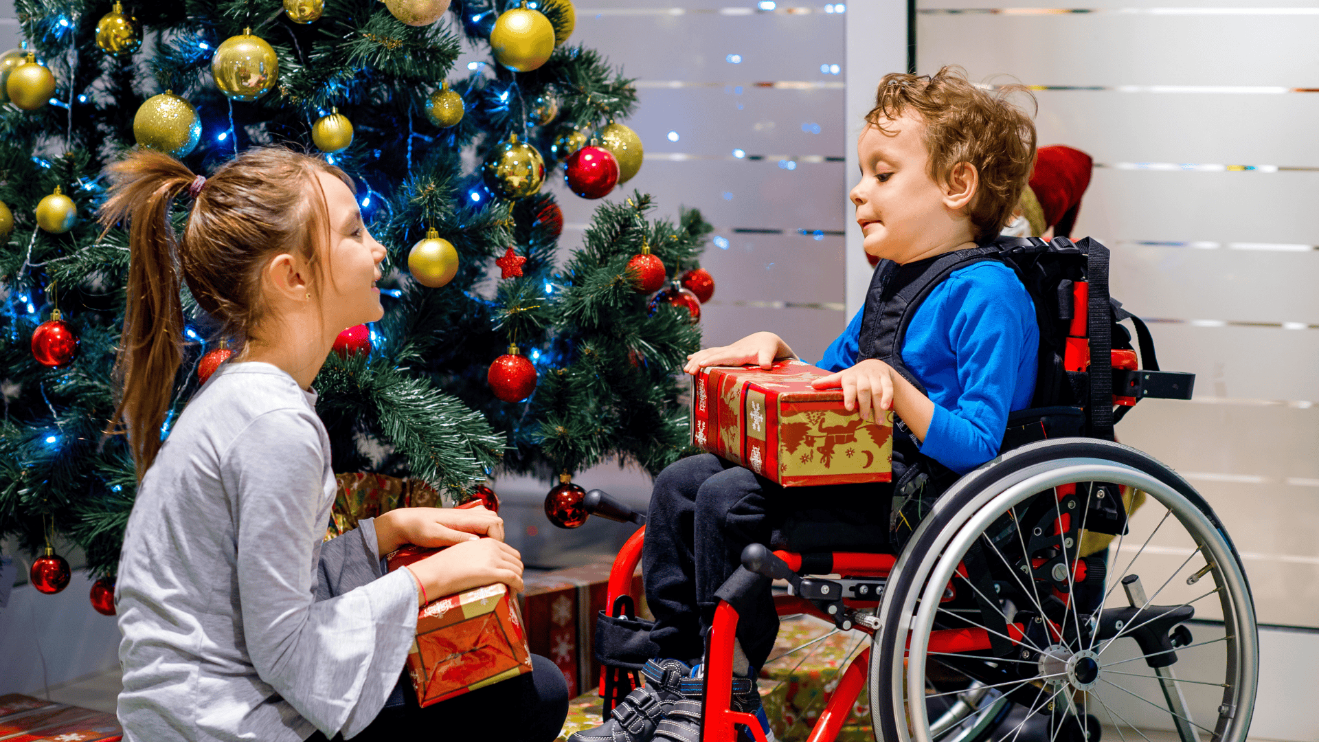 Two children, one in a wheelchair, exchange gifts in front of a Christmas tree.